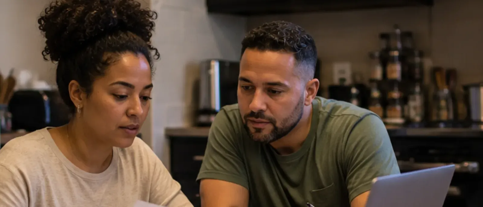 A couple sitting together at a kitchen table, going over a paper statement beside an open laptop and a mug of coffee.