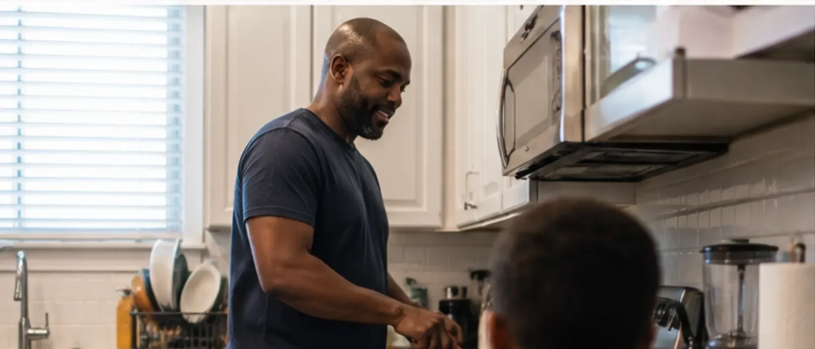 A father cooking breakfast at the stove in a home kitchen while his child, seen from behind, waits nearby.